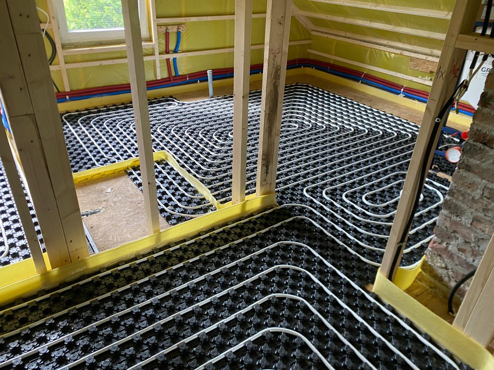 Widok na instalację ogrzewania podłogowego w trakcie montażu, rury w systemie rozprowadzonym na czarnej macie, widoczne elementy konstrukcji drewnianej poddasza i izolacja żółtą folią.