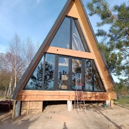 Profil Sp. z.o.o. - Nowoczesny dom w kształcie litery A z dużymi przeszkleniami, drewnianą fasadą i czerwoną drabiną opartą o ścianę, w tle drzewa i błękitne niebo.