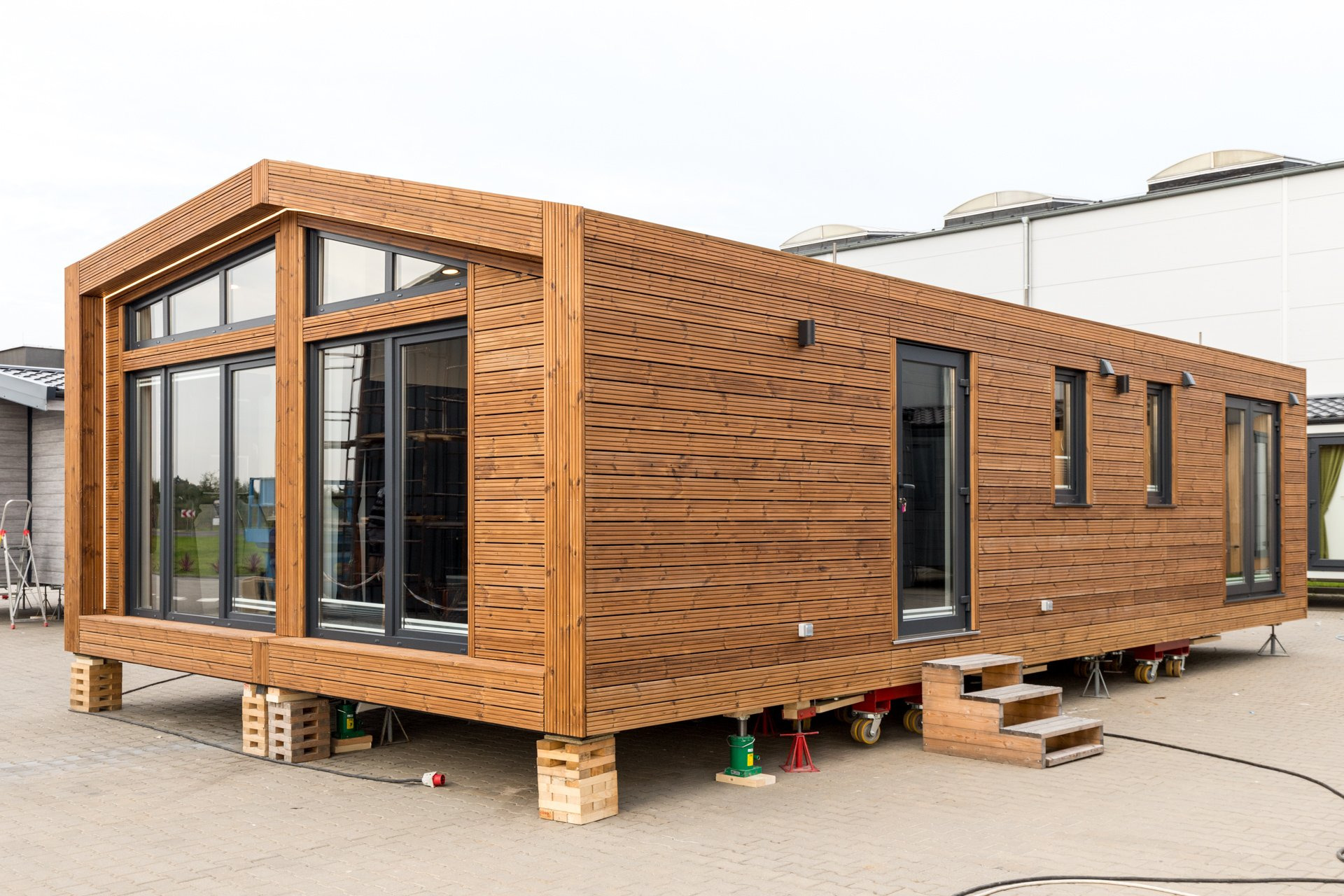 Gotowy modułowy dom z drewnianą elewacją, ustawiony na tymczasowych podporach przed montażem na fundamencie, z widocznymi oknami i drzwiami.