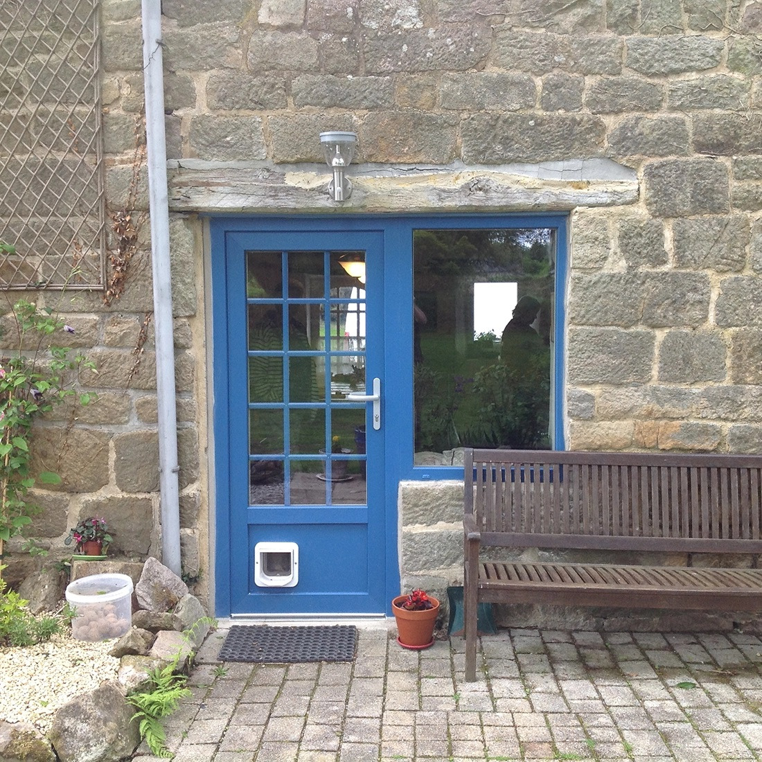 Błękitne drzwi zewnętrzne z okienkiem i kocim przejściem, wkomponowane w kamienną elewację domu w Szczecinku, obok drewniana ławka i doniczki z kwiatami.