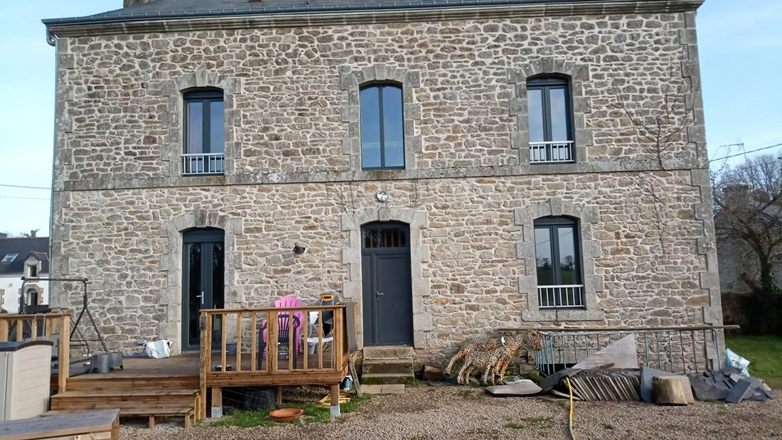 Fasada kamiennego domu z nowymi oknami i drzwiami w kolorze antracytowym, widoczne dekoracje ogrodowe i taras z meblami.