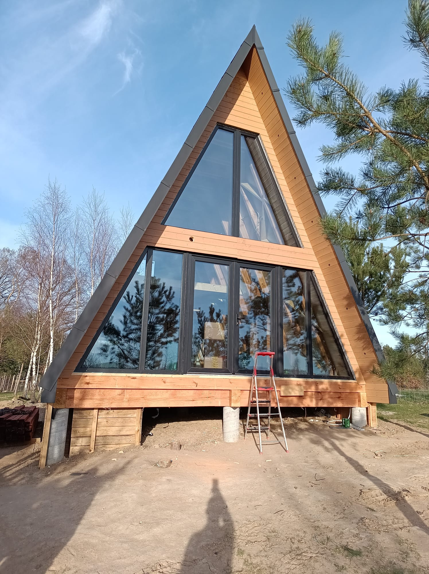 Nowoczesny dom w kształcie litery A z dużymi przeszkleniami, drewnianą fasadą i czerwoną drabiną opartą o ścianę, w tle drzewa i błękitne niebo.