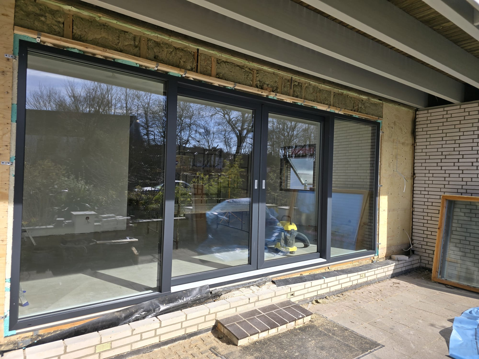 Montaż trzyskrzydłowych okien przesuwnych PCV w surowym stanie, widoczna izolacja ściany i narzędzia pracy.