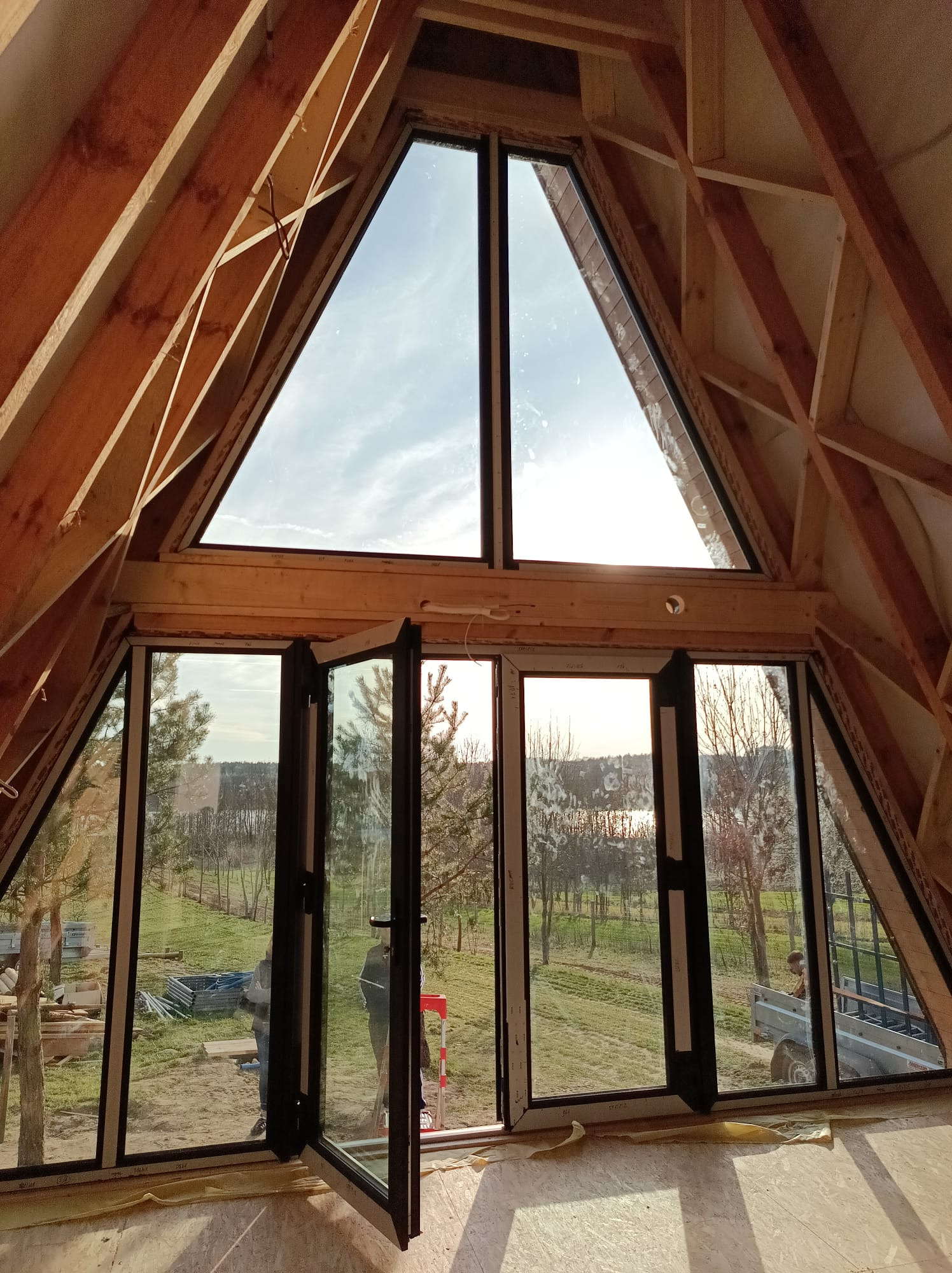 Montaż okien w domu w stanie surowym zamkniętym, widok z wnętrza na szkielet dachu i otwarty krajobraz za oknami.