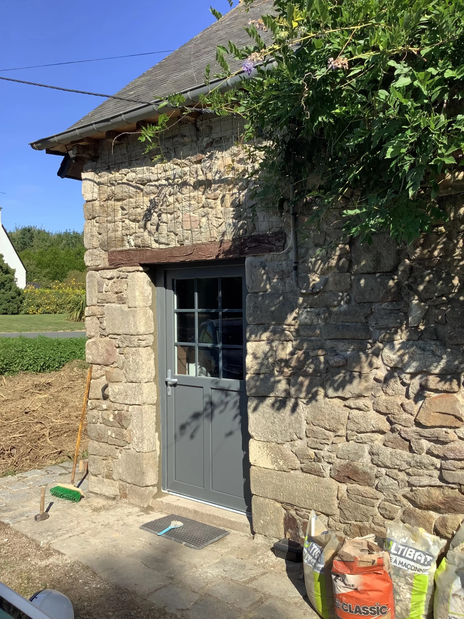 Szara, przeszklona, nowoczesna stolarka drzwiowa w kamiennym budynku z elementami roślinności, widoczne worki z zaprawą murarską, szczotka i młotek.