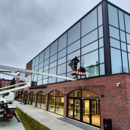 WITEK Łukasz Witkowski - Dwóch pracowników w koszu podnośnika na tle ceglanego budynku z dużą szklaną fasadą, myjących okna na wysokości, widoczny fragment chodnika i trawnika oraz zaparkowany samochód z podnośnikiem.