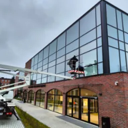 Dwóch pracowników w koszu podnośnika na tle ceglanego budynku z dużą szklaną fasadą, myjących okna na wysokości, widoczny fragment chodnika i trawnika oraz zaparkowany samochód z podnośnikiem.
