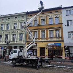 WITEK Łukasz Witkowski - Pracownik w koszu podnośnika na wysokości dachu kamienicy podczas prac na zewnątrz w pochmurny dzień, widoczne zabezpieczenia taśmą ostrzegawczą na chodniku.