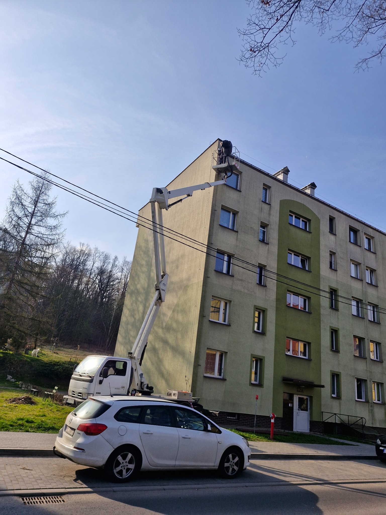 Pracownik w koszu podnośnika na wysokości dachu budynku wielorodzinnego, potencjalnie podczas prac konserwacyjnych lub naprawczych.