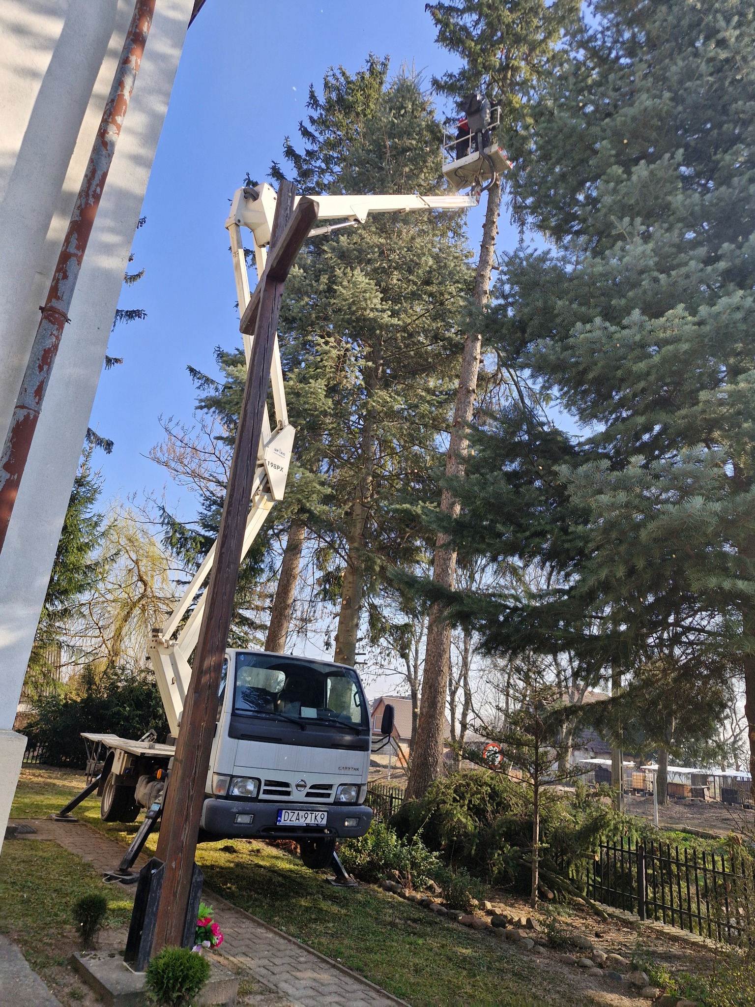 Pracownik w koszu podnośnika tnie wysokie drzewo iglaste przy budynku kościoła, obok drewniany krzyż i zaparkowany samochód z podnośnikiem koszowym.