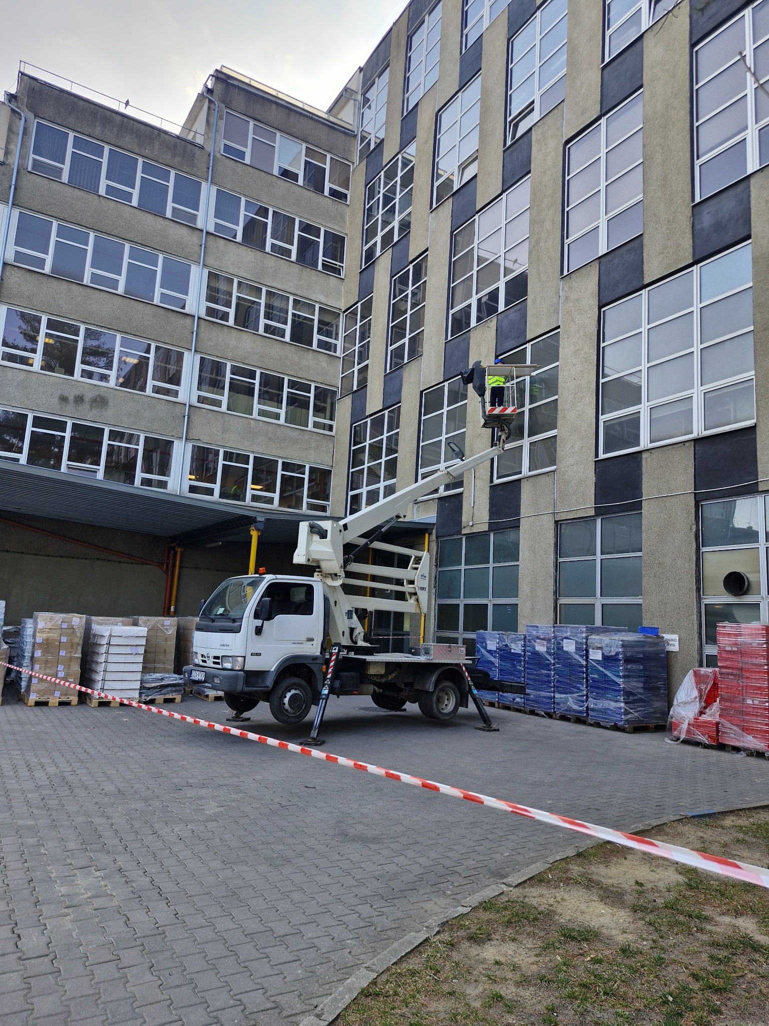 Pracownik w koszu podnośnika na tle budynku z szarą elewacją i licznymi oknami. Na dole palety z materiałami budowlanymi i taśma ostrzegawcza.