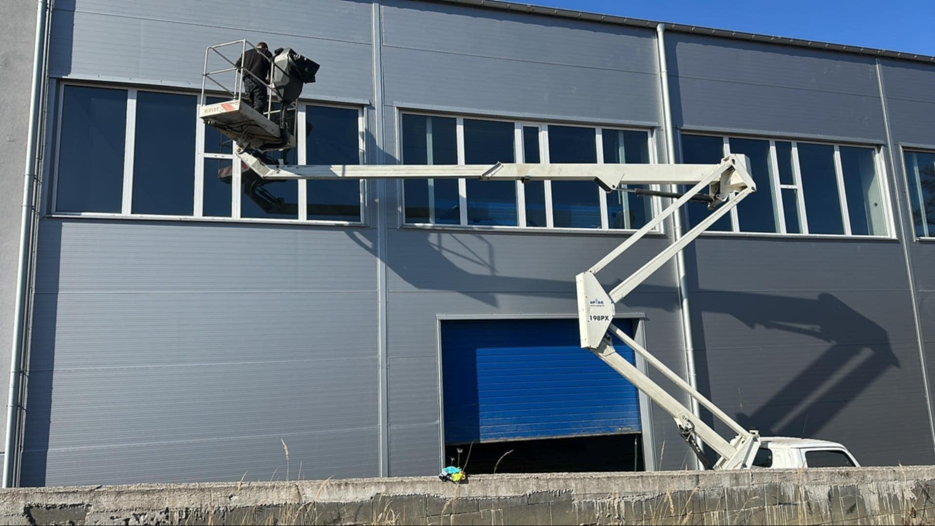 Dwóch pracowników w koszu podnośnika na tle szarej hali z rzędami okien, niebieską bramą i częściowo widocznym pojazdem