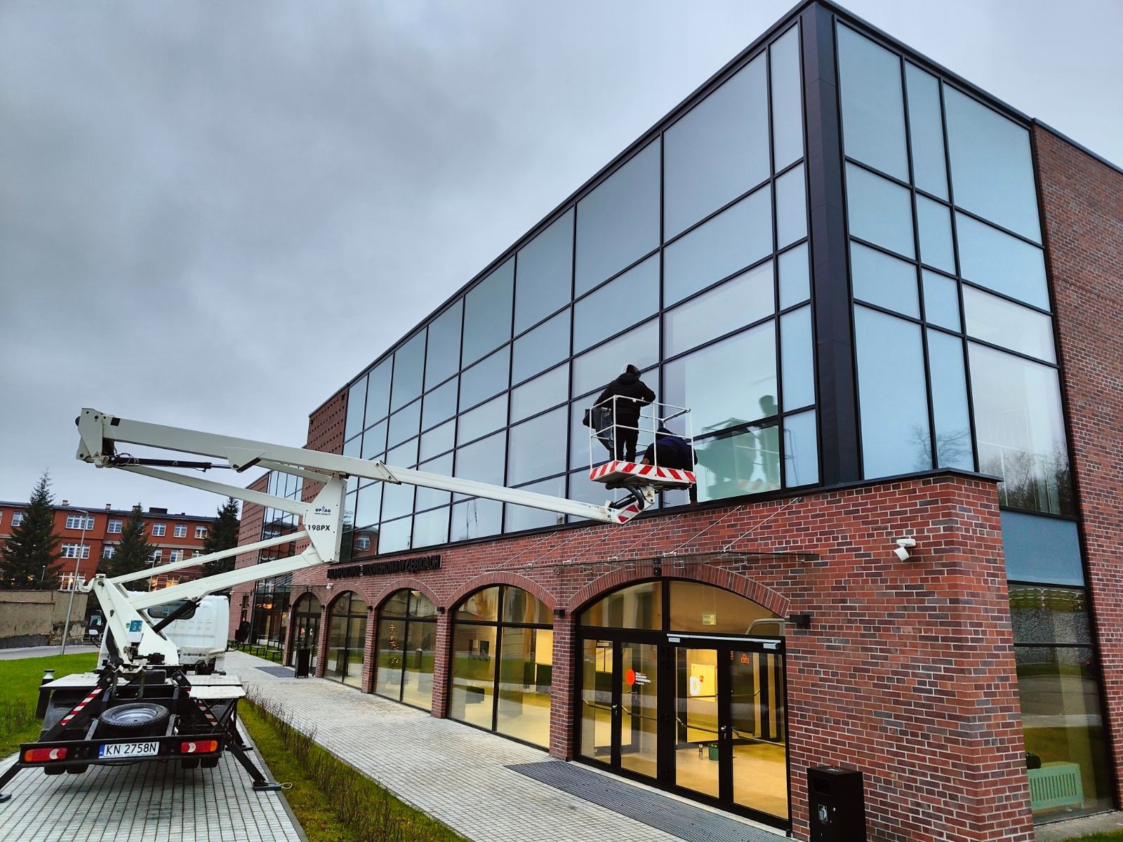 Dwóch pracowników w koszu podnośnika na tle ceglanego budynku z dużą szklaną fasadą, myjących okna na wysokości, widoczny fragment chodnika i trawnika oraz zaparkowany samochód z podnośnikiem.