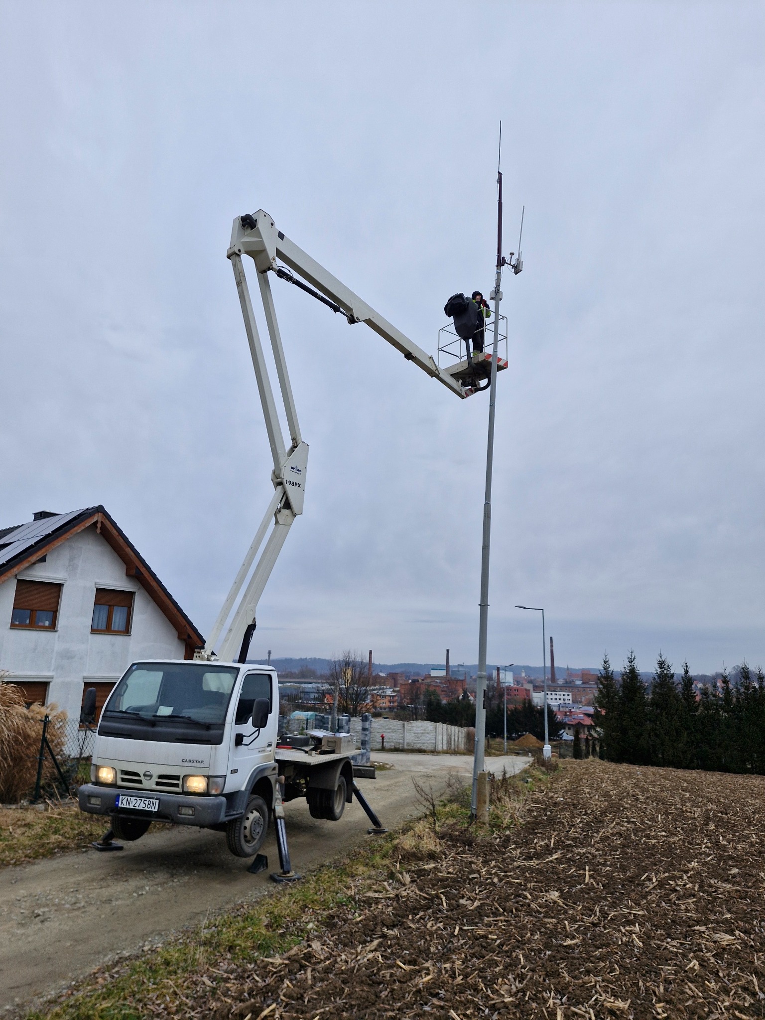 Pracownik na podnośniku koszowym montuje antenę na słupie telekomunikacyjnym; w tle dom z panelami słonecznymi i zabudowania miejskie.