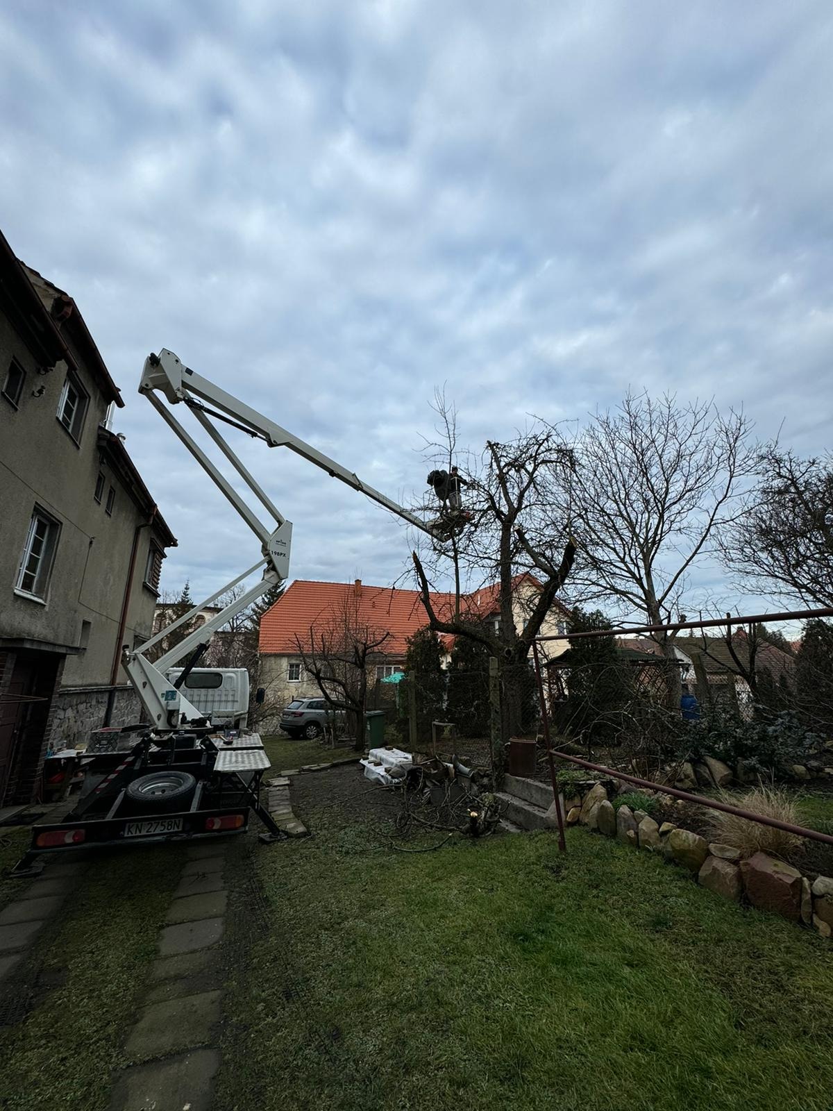 Pracownik w koszu podnośnika tnący gałęzie drzewa w ogrodzie, widoczny budynek mieszkalny i fragment podnośnika koszowego na tle pochmurnego nieba.