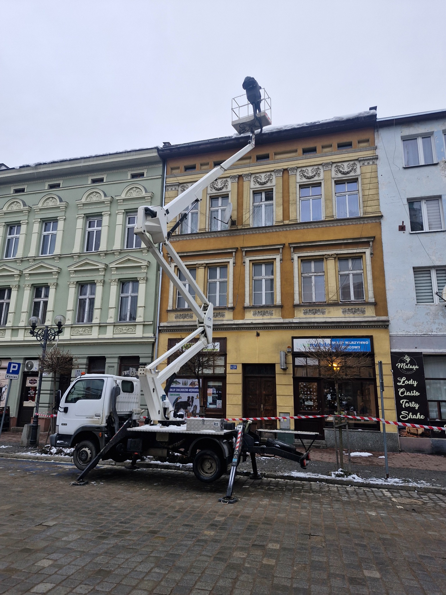 Pracownik w koszu podnośnika na wysokości dachu kamienicy podczas prac na zewnątrz w pochmurny dzień, widoczne zabezpieczenia taśmą ostrzegawczą na chodniku.