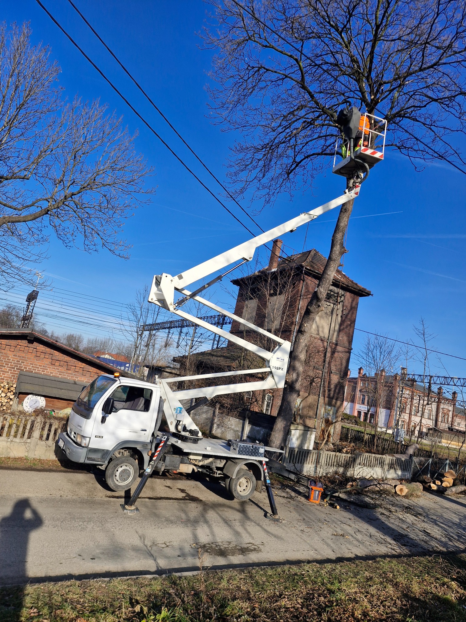 Pracownik w pomarańczowej kamizelce na podnośniku koszowym przycina gałęzie drzewa, w tle budynki i niebo z liniami energetycznymi.