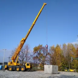 Żółty dźwig samochodowy ZAGAJEWSKI podnosi betonowy blok na placu budowy, widoczny przenośny toi-toi i drzewa z jesiennymi liśćmi w tle.