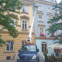 EHC Karol Zagajewski sp. k. - Podnośnik koszowy na tle kamienic w trakcie pracy, pracownik w koszu na wysokości okien drugiego piętra, niebieski samochód bazowy z numerem telefonu 502 602 577.