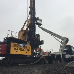 EHC Karol Zagajewski sp. k. - Żółto-czerwona wiertnica Aarsleff na gąsienicach, wspierana przez podnośnik koszowy na tle szarego nieba i ziemi.