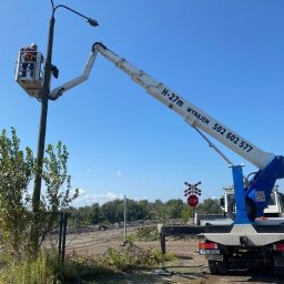 EHC Karol Zagajewski sp. k. - Podnośnik koszowy H-27m z numerem telefonu wynajmu, podnosi pracownika do słupa oświetleniowego na tle błękitnego nieba i zieleni.