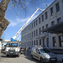 Podnośnik koszowy H-27m na podwoziu MAN, z wysięgnikiem sięgającym dachu budynku, zaparkowany na chodniku obok drzewa i zaparkowanego srebrnego vana.