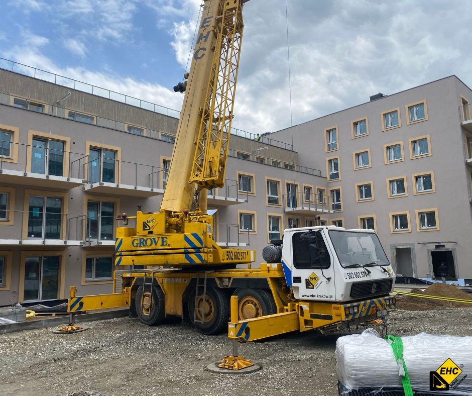 Żółty dźwig samochodowy Grove z wysuniętym ramieniem na placu budowy wielopiętrowego budynku mieszkalnego, widoczne podpory i numer telefonu na boku pojazdu.