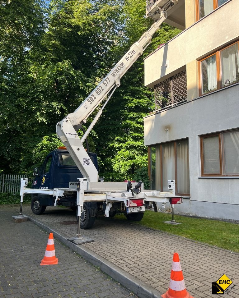 Podnośnik koszowy Ruthmann H-22m ustawiony na chodniku z rozłożonymi podporami, z widocznym numerem telefonu i tablicą rejestracyjną K4 EHC, obok budynku z oknami i balkonem
