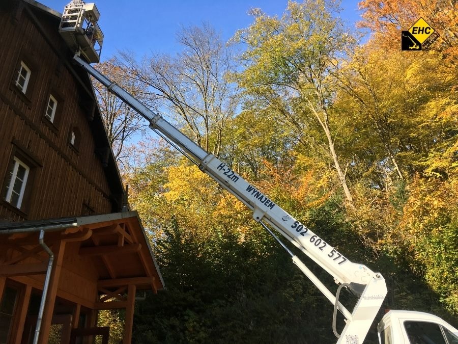 Biały podnośnik koszowy H-22m na tle drewnianego budynku i jesiennego lasu, z widocznym numerem telefonu kontaktowego na ramieniu oraz logo firmy w prawym górnym rogu.