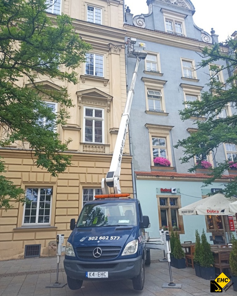 Podnośnik koszowy na tle kamienic w trakcie pracy, pracownik w koszu na wysokości okien drugiego piętra, niebieski samochód bazowy z numerem telefonu 502 602 577.