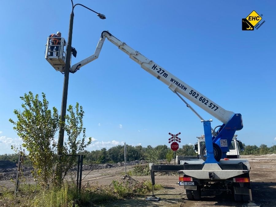 Podnośnik koszowy H-27m z numerem telefonu wynajmu, podnosi pracownika do słupa oświetleniowego na tle błękitnego nieba i zieleni.