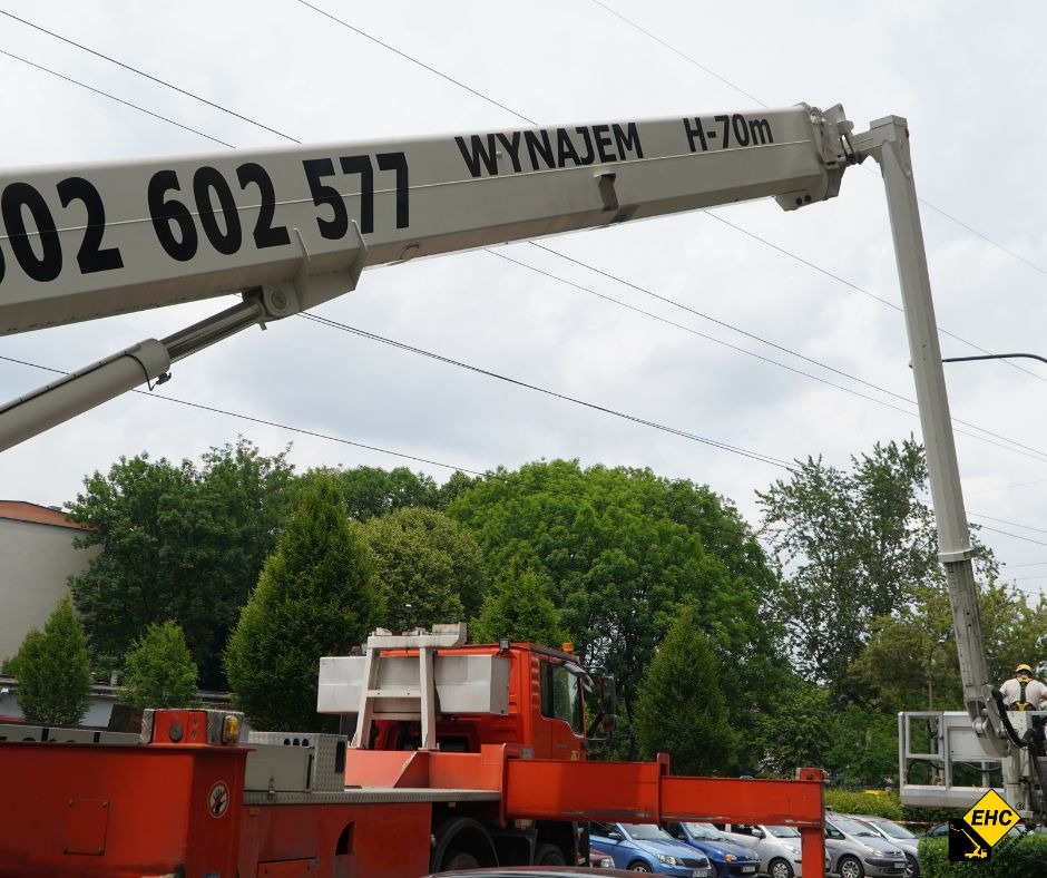 Pomarańczowy podnośnik koszowy z wysięgnikiem z napisem 'WYNAJEM H-70m' i numerem telefonu, na tle zieleni drzew i pochmurnego nieba, z pracownikami w koszu na wysokości.