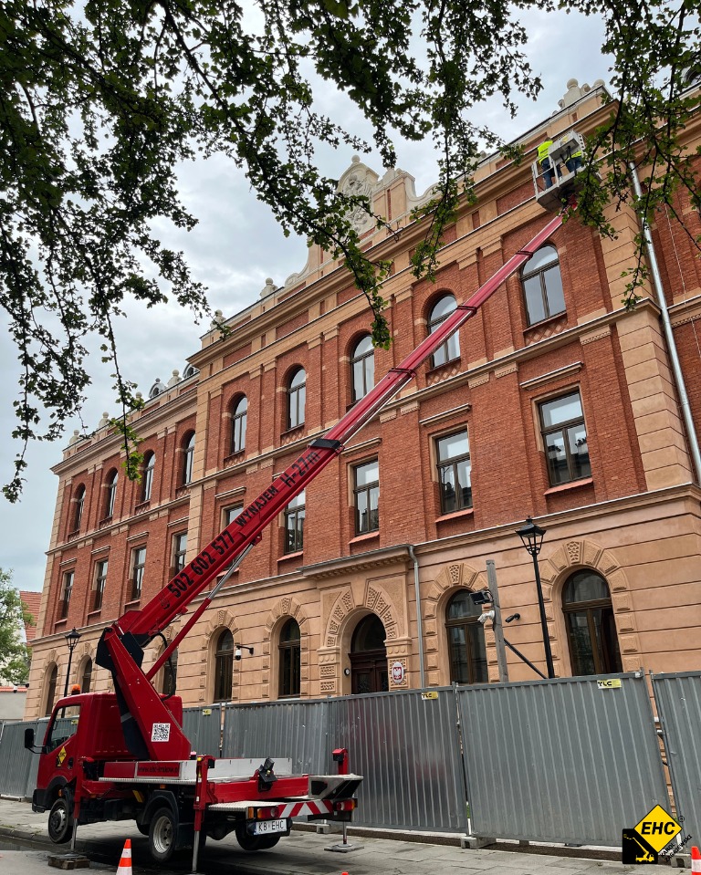 Czerwony podnośnik koszowy na tle ceglanego budynku z ozdobnymi elementami architektonicznymi, pracownicy w koszu na wysokości przy elewacji, widoczne zabezpieczenia i drzewa w kadrze.