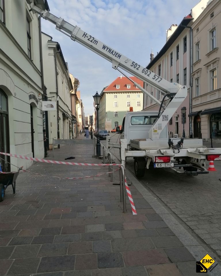 Podnośnik koszowy H-22m na podwoziu samochodu dostawczego, zaparkowany na brukowanej ulicy w centrum miasta, zabezpieczony taśmami ostrzegawczymi.