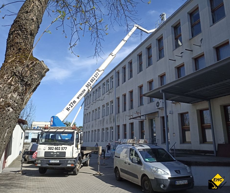 Podnośnik koszowy H-27m na podwoziu MAN, z wysięgnikiem sięgającym dachu budynku, zaparkowany na chodniku obok drzewa i zaparkowanego srebrnego vana.