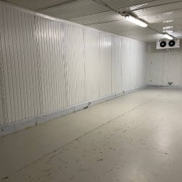 V&J GROUP SPÓŁKA Z OGRANICZONĄ ODPOWIEDZIALNOŚCIĄ - Pusta, biała hala magazynowa z panelami ściennymi, betonową podłogą z widocznymi śladami użytkowania i dwoma klimatyzatorami pod sufitem.