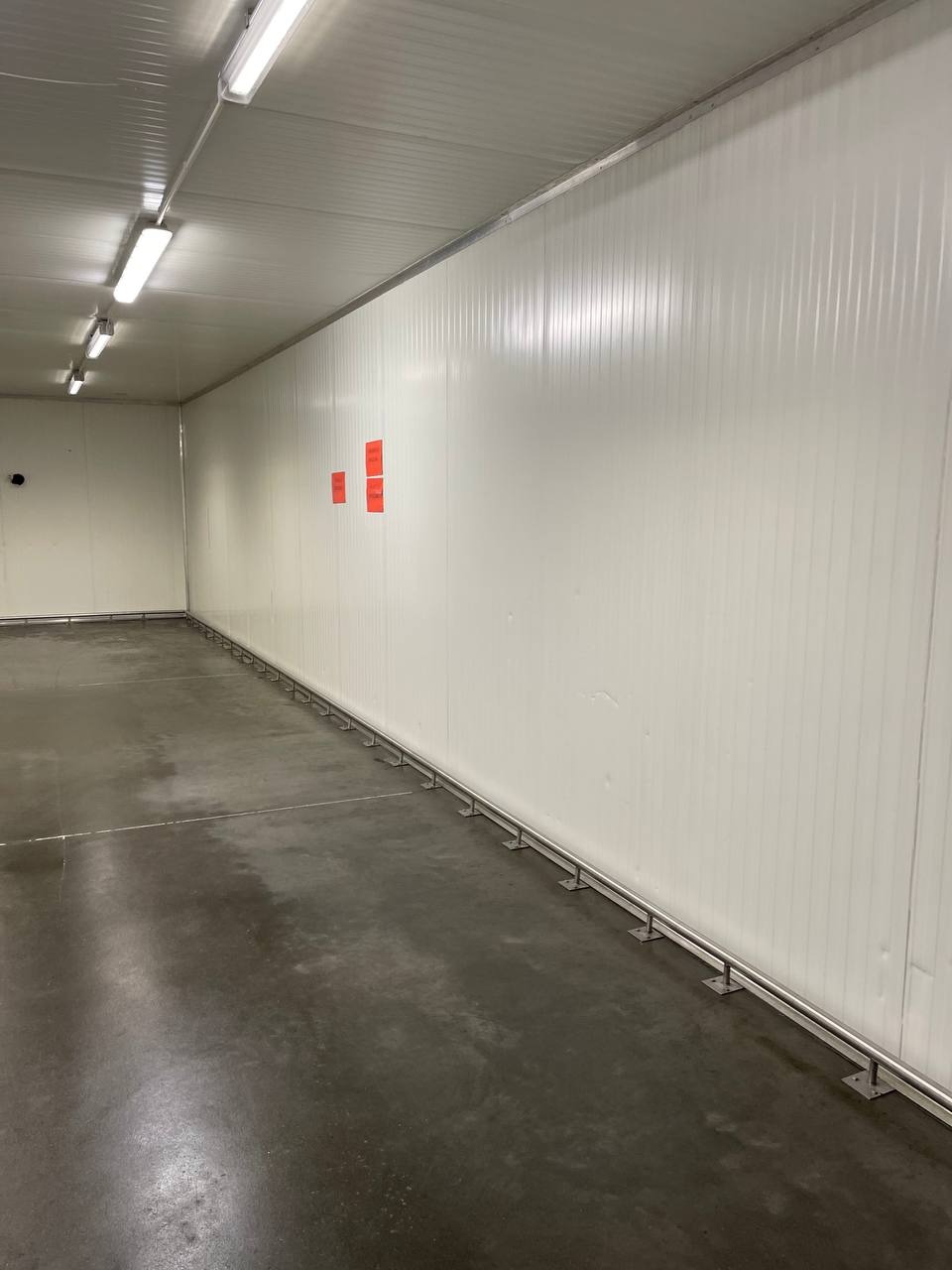 Pusta hala z białymi panelami ściennymi i betonową podłogą, widoczne rury przy ścianie i pomarańczowe naklejki na panelach.