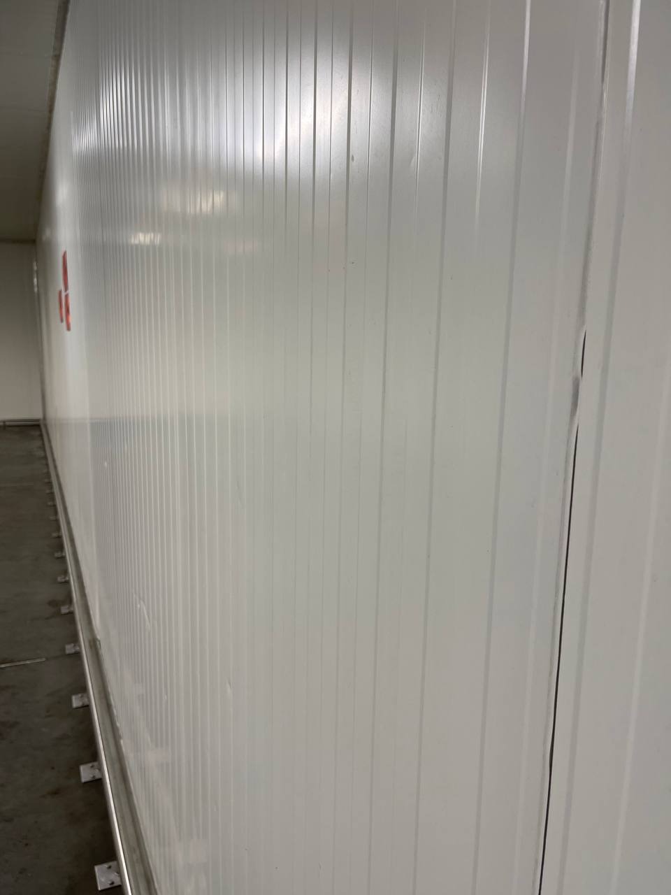Długa, biała, pofałdowana ściana w pomieszczeniu przemysłowym, widziana pod kątem, z betonową podłogą i fragmentem sufitu.