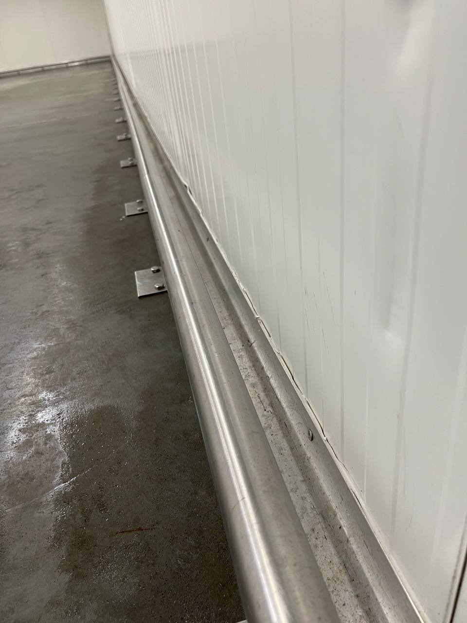 Wnętrze hali produkcyjnej lub magazynowej z panelami ściennymi i zabezpieczeniem przypodłogowym z metalowych rur, widoczne ślady użytkowania na podłodze i ścianach.