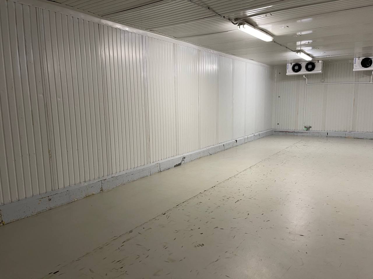 Pusta, biała hala magazynowa z panelami ściennymi, betonową podłogą z widocznymi śladami użytkowania i dwoma klimatyzatorami pod sufitem.