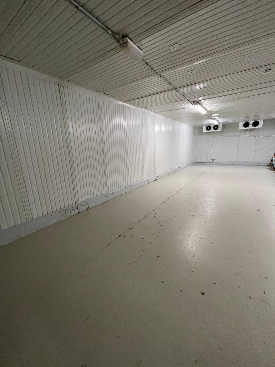Pusta hala magazynowa z panelami ściennymi i śladami zabrudzeń na podłodze, widoczne urządzenia chłodnicze pod sufitem.