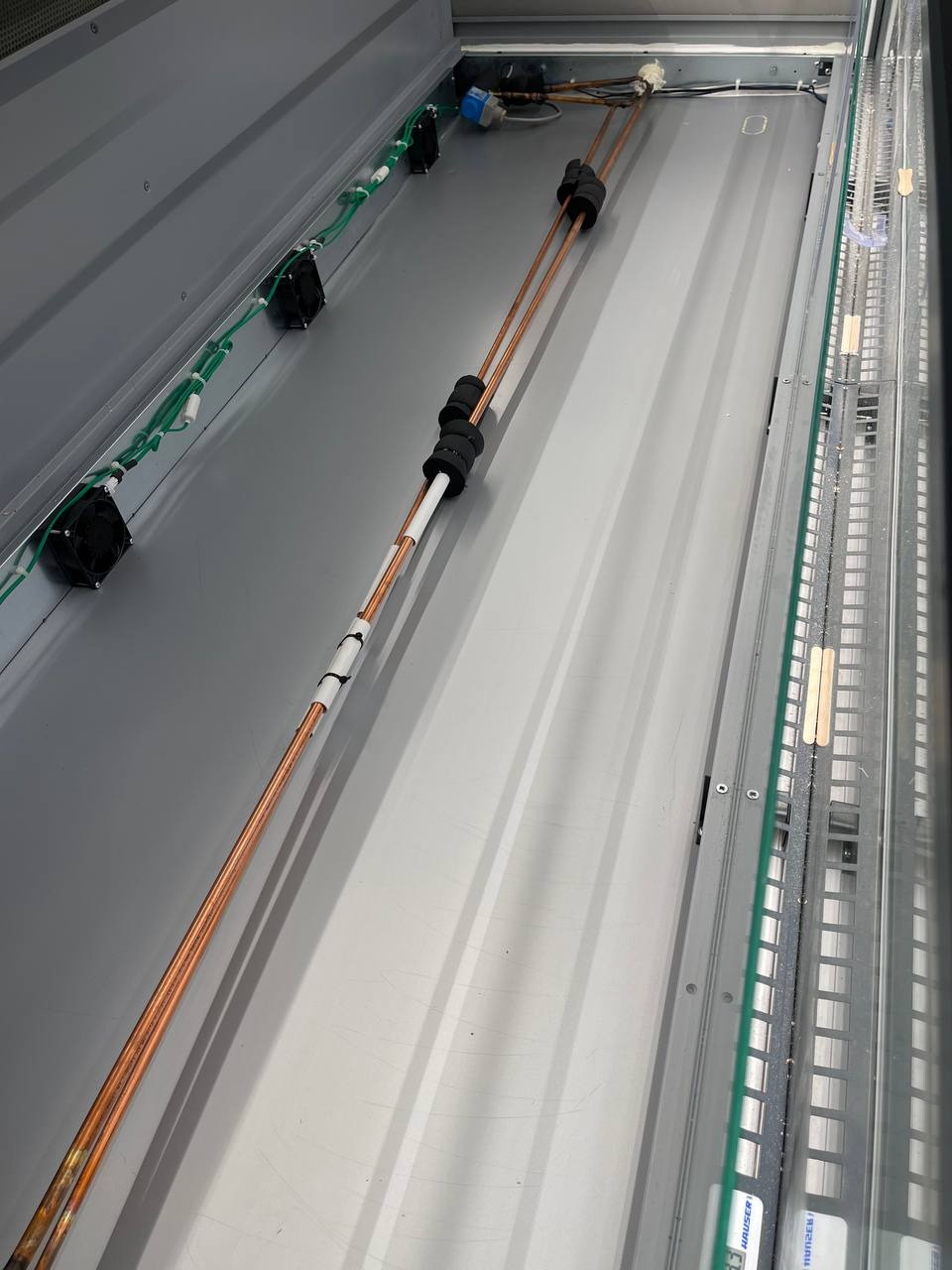 Wnętrze urządzenia chłodniczego z widocznymi miedzianymi rurkami, czarnymi izolatorami i zielonymi przewodami w szarej obudowie.