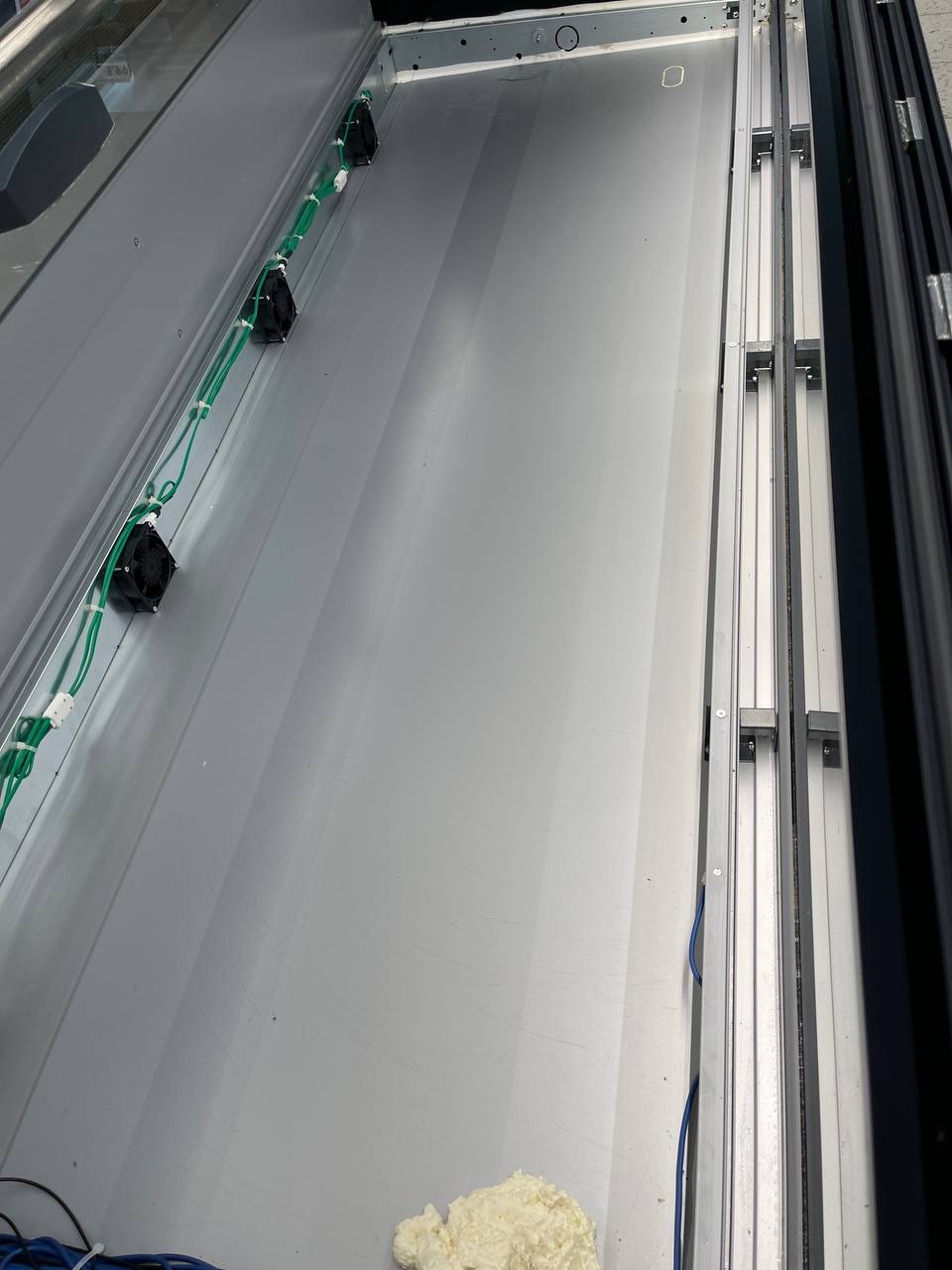 Wnętrze pustej, metalowej skrzyni z widocznymi przewodami i wentylatorami po jednej stronie oraz aluminiowymi profilami po drugiej, z resztkami pianki montażowej na dnie.