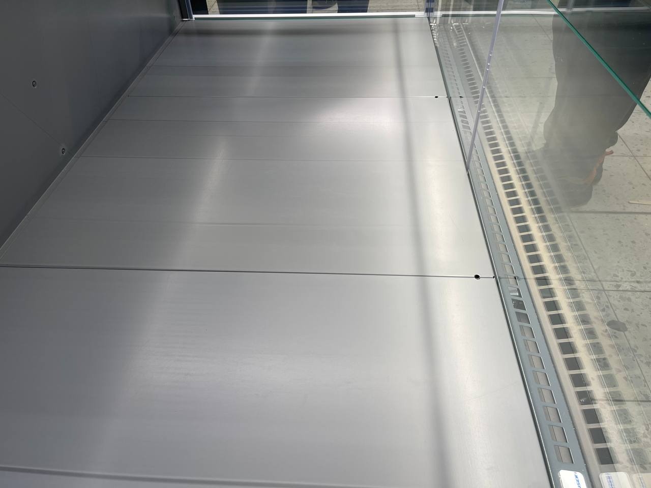 Wnętrze chłodniczej witryny sklepowej z metalowymi panelami i szklaną szybą, widoczne odbicie butów osoby fotografującej.