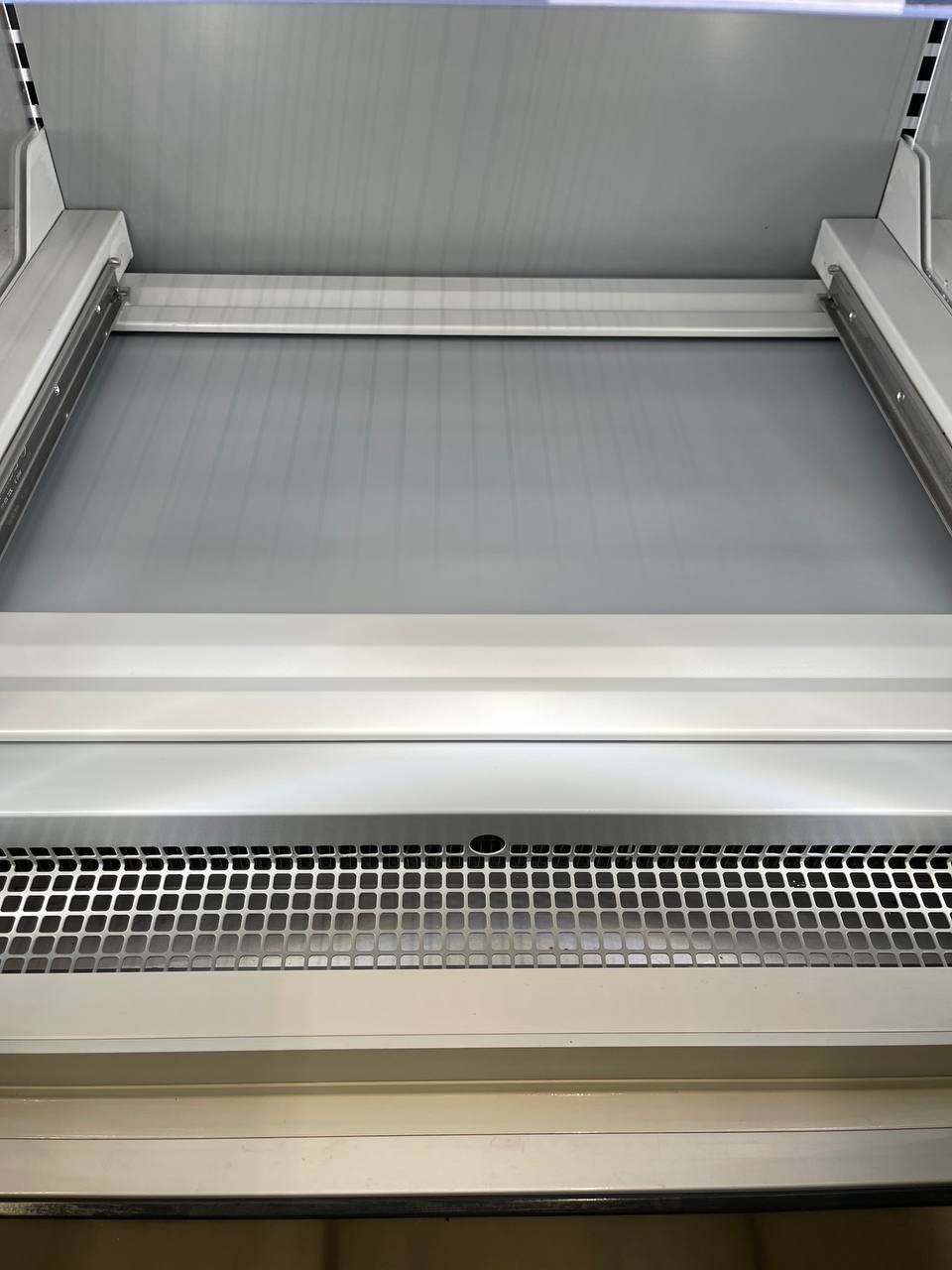 Wnętrze czystej, pustej witryny chłodniczej z metalową kratką wentylacyjną, oświetleniem LED i gładkimi, białymi powierzchniami.