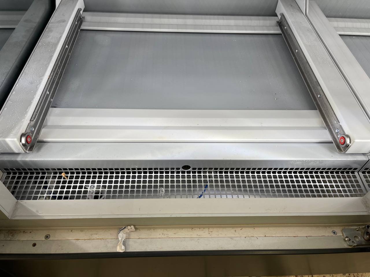 Wnętrze szafki lub urządzenia chłodniczego z metalową kratką wentylacyjną, widoczne zabrudzenia i resztki w dolnej części.