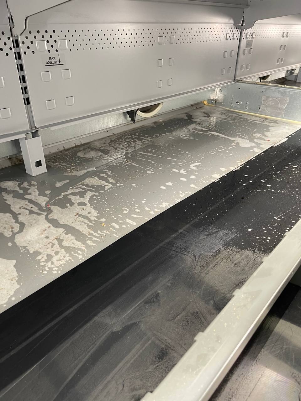 Wnętrze przemysłowej maszyny z widocznymi śladami czyszczenia, mokra podłoga z pianą i resztkami brudu, czarna gumowa listwa. Widoczna naklejka z maksymalnym obciążeniem 300kg/m2.