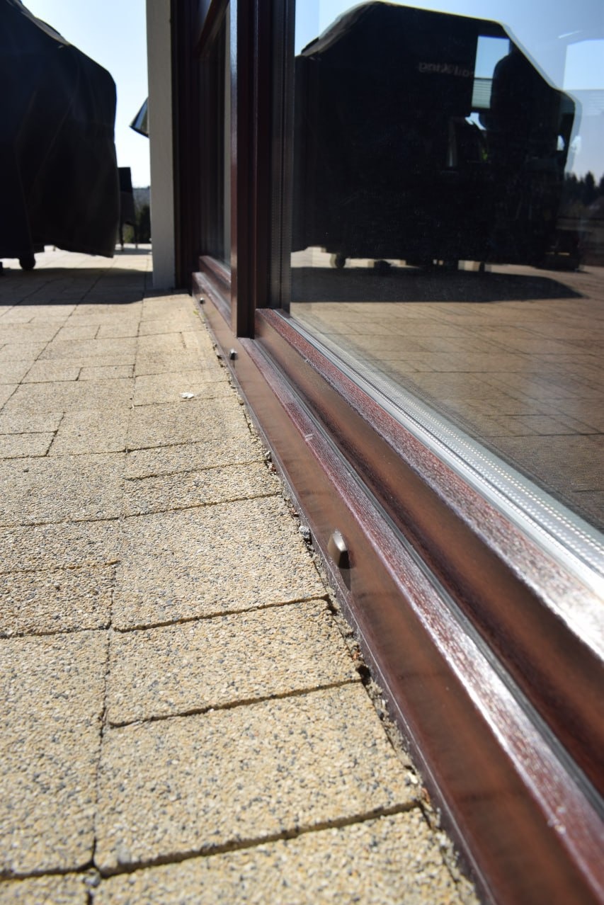 Dolna część brązowych, przesuwnych drzwi balkonowych z widocznymi zabrudzeniami w szczelinach, na tle kostki brukowej i odbicia otoczenia w szybie.