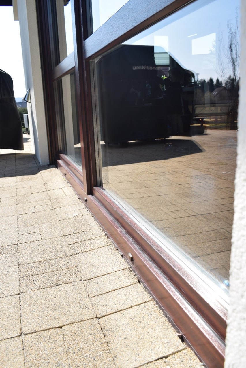 Dolna część okna przesuwnego z brązową ramą, widoczna z zewnątrz na tle kostki brukowej; w szybie odbija się budynek i pojazd.