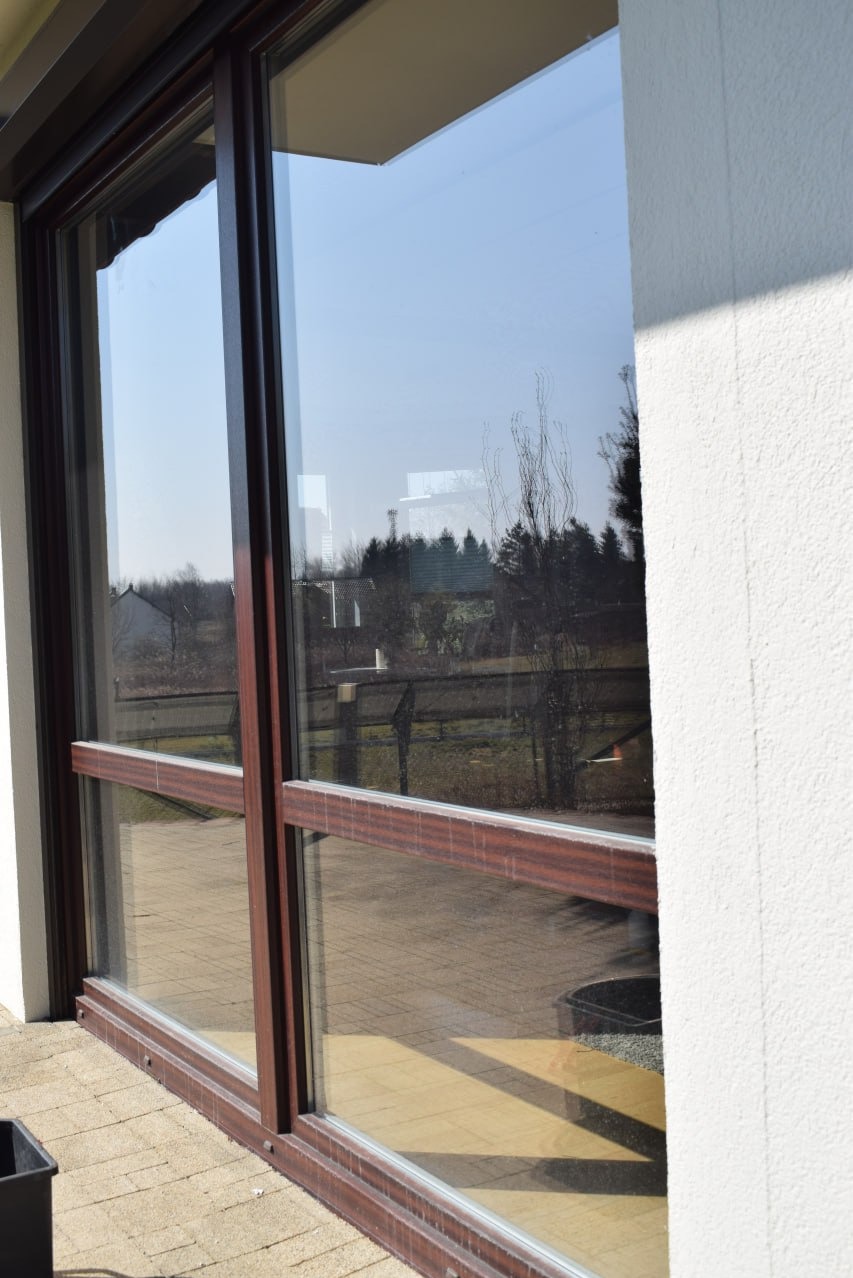 Czyste okno tarasowe z brązowymi ramami w Krakowie, odbijające drzewa i budynki. Widoczny fragment białej ściany i kostka brukowa.